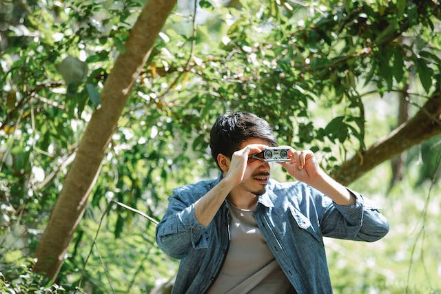 A man uses a small pair of binoculars in the woods. 
