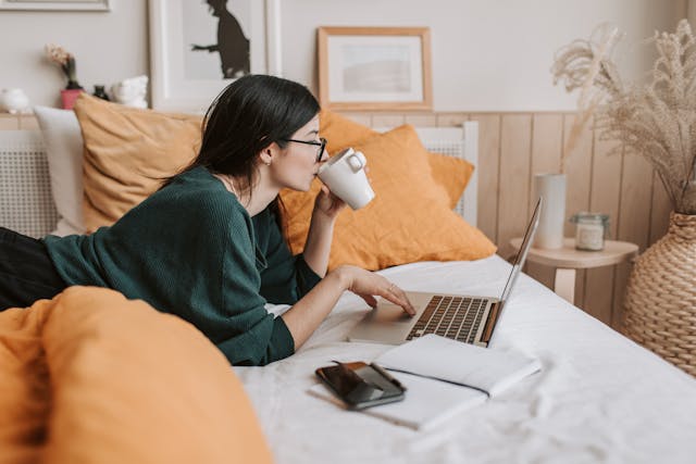 Een vrouw ligt op haar buik op bed en drinkt uit een beker terwijl ze video's bekijkt op haar laptop. 