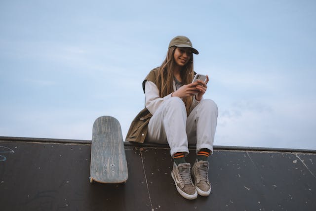 Una giovane donna si riposa accanto al suo skateboard e guarda TikToks sul suo telefono. 