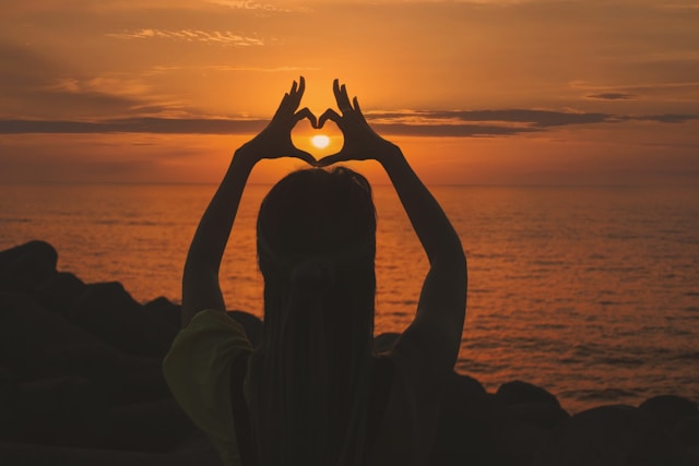 A woman’s silhouette frames the setting sun inside her heart-shaped fingers. 