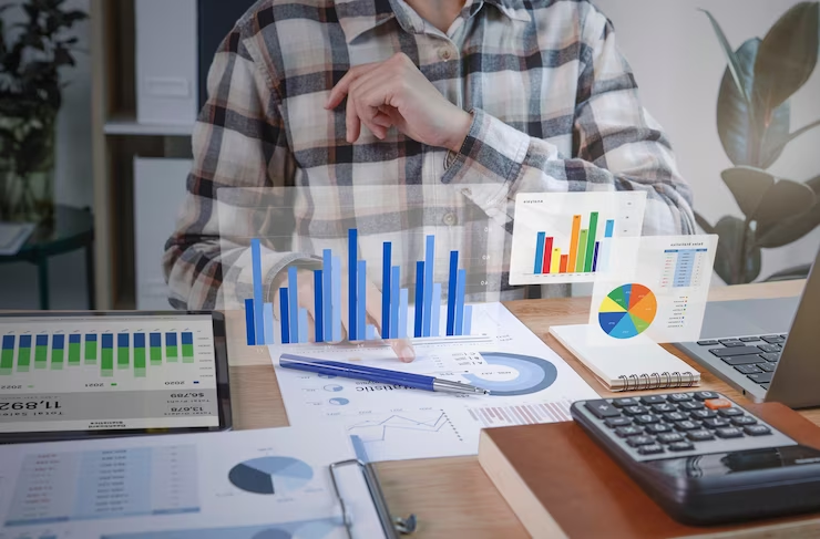 A person sits on a desk with a laptop, tab, papers, and floating graphs.