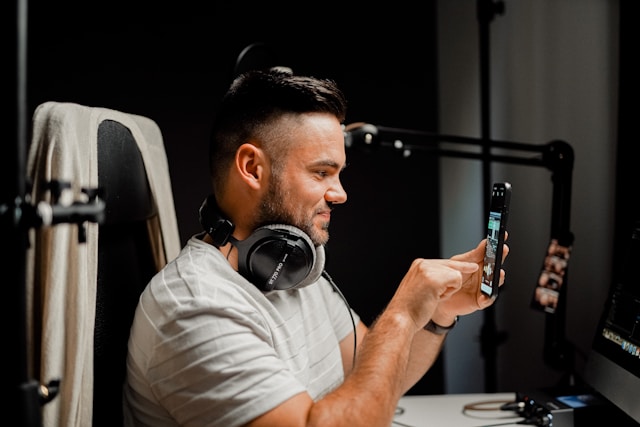A man in a white T-shirt with headphones records a video on a smartphone.