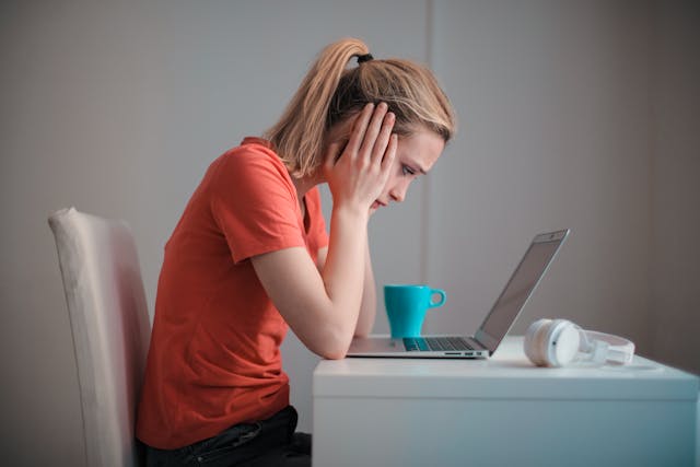 Een vrouw kijkt bedenkelijk terwijl ze met haar ellebogen op tafel en haar gezicht tussen haar handen naar haar laptop staart.