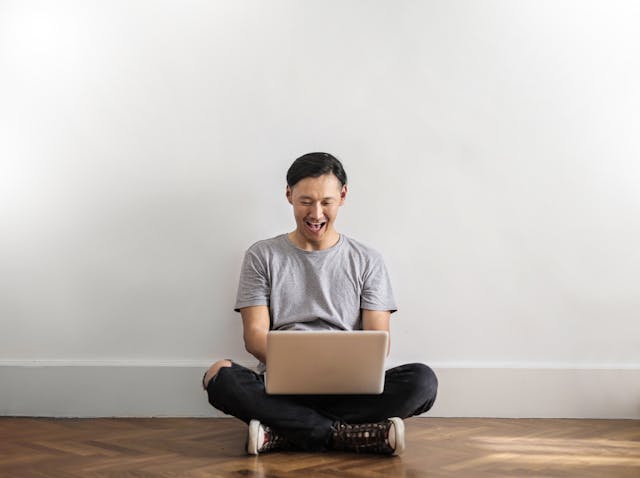 A person sits on the floor and laughs while browsing on their laptop.