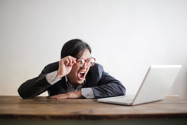 A man holds his glasses and yells at his laptop. 