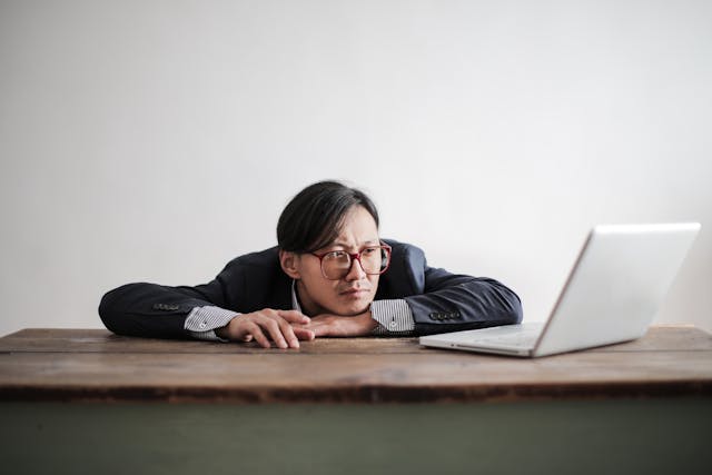 Een man laat zijn kin rusten en tikt ongeduldig met zijn vingers op een tafel voor zijn laptop.