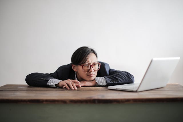 A man rests his chin and impatiently taps his fingers on a table in front of his laptop.