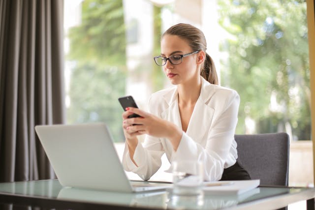 一位女性专业人士坐在笔记本电脑前用手机打字。 