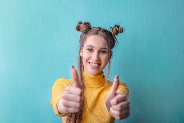 O tânără cu un pulover galben dă două degete în sus. 