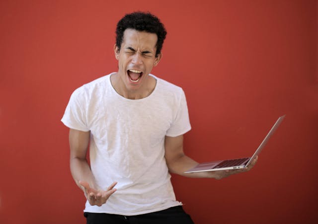 Un homme tient son ordinateur portable en équilibre sur une main tout en hurlant de frustration.