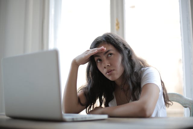 A woman rubs her temples with her hand as she sits frustrated in front of her laptop. 