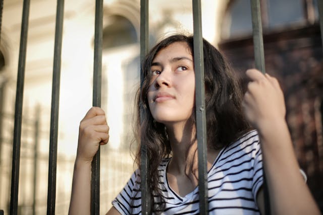A girl grabs the metal bars of a tall fence. 
