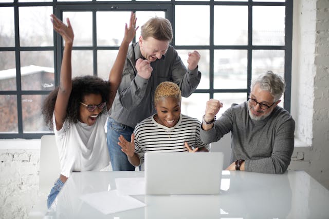 A group of cheering coworkers gather around a laptop.