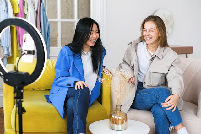 Two women sit on armchairs in front of a ring light and smartphone and share a laugh while they host a TikTok LIVE. 