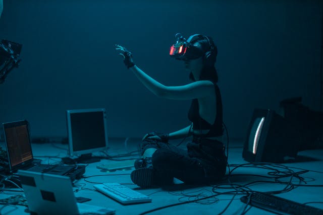 A woman wears a VR headset as she sits on the floor in front of several computers.