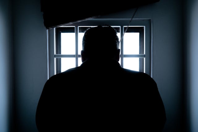 The silhouette of a man standing in shadow in front of a window with bars.