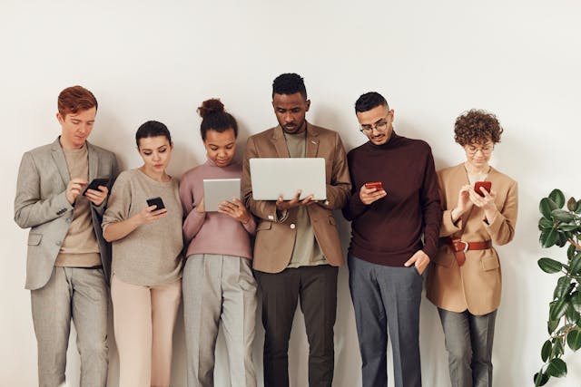 A group of young professionals checks their own devices as they stand against a wall. 
