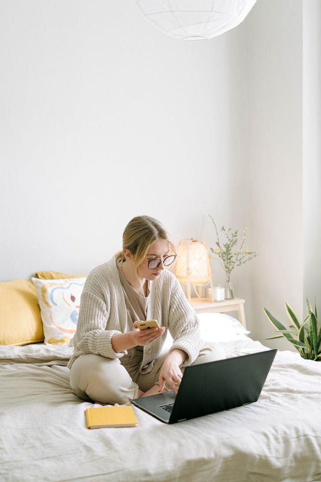 A woman holds a phone while she types on her laptop.
