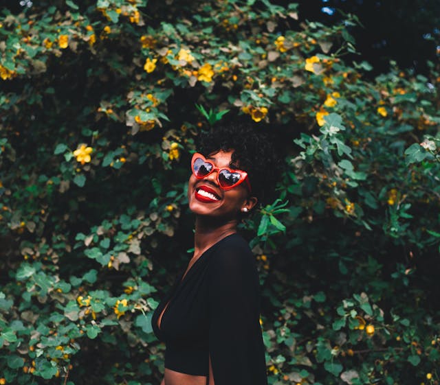 Eine Frau steht lächelnd vor einem Busch und trägt eine rote Sonnenbrille.