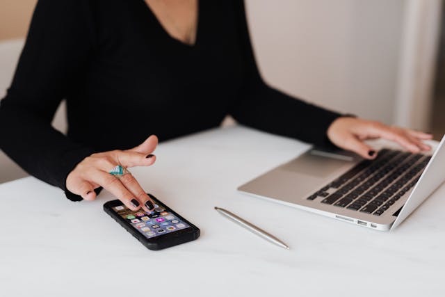 Uma mulher faz uma pausa no trabalho em seu laptop para verificar as notificações em seu telefone. 