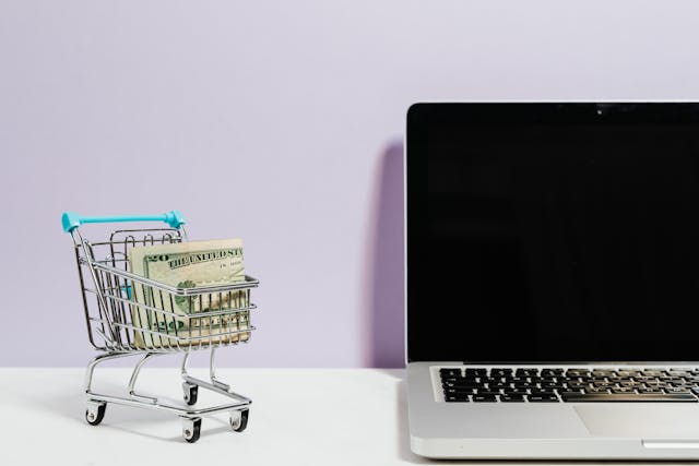 A tiny shopping cart with dollar bills in it stands beside a laptop.