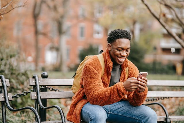 A man sits at a park bench and smiles while he looks at his phone. 
