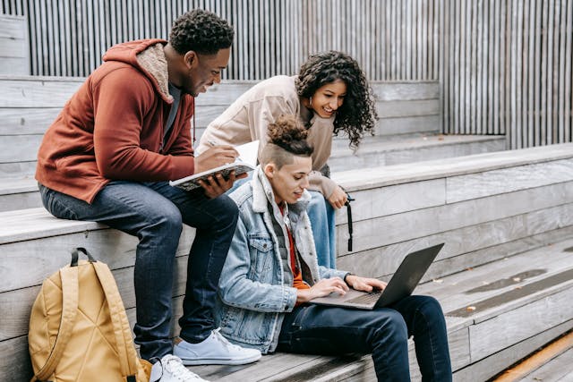 A group of friends sits on wooden benches and shares a laptop to watch videos. 
