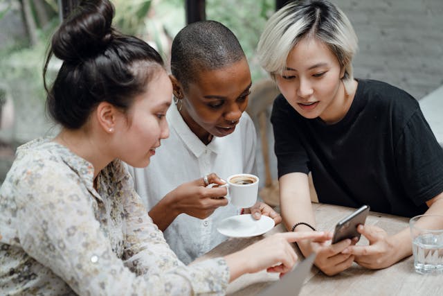 Three female friends sit at an outdoor cafe and use a phone to check social media updates. 
