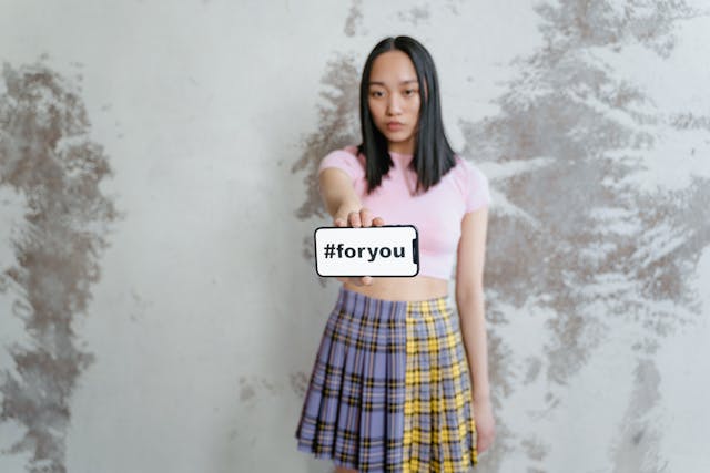 A young woman holds up a phone that shows the hashtag “#foryou” on its screen.

