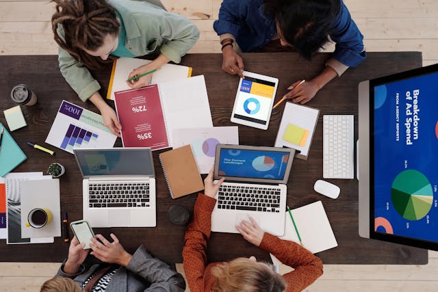 A group of young professionals discusses advertising strategies with laptops, tablets, and graphs in front of them.