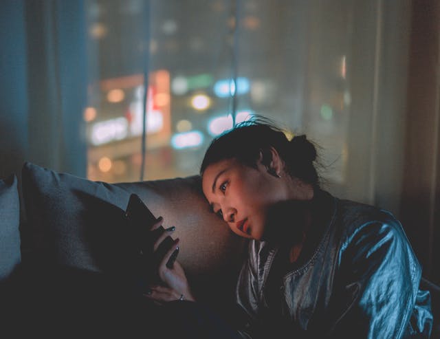 Dans une chambre sombre, une femme pose sa tête sur un oreiller et consulte son téléphone. 