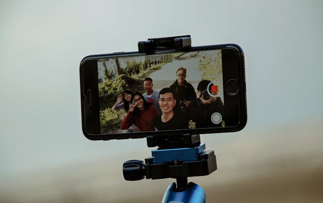 Um smartphone preto em um tripé registra um grupo de amigos sorrindo ao ar livre.