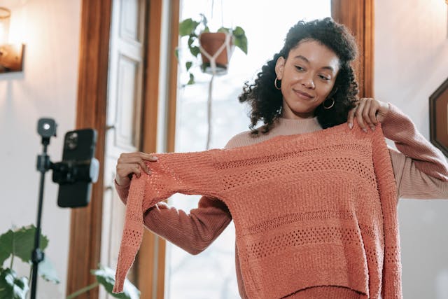 A woman holds up an orange knit sweater in front of her smartphone to show her LIVE audience.