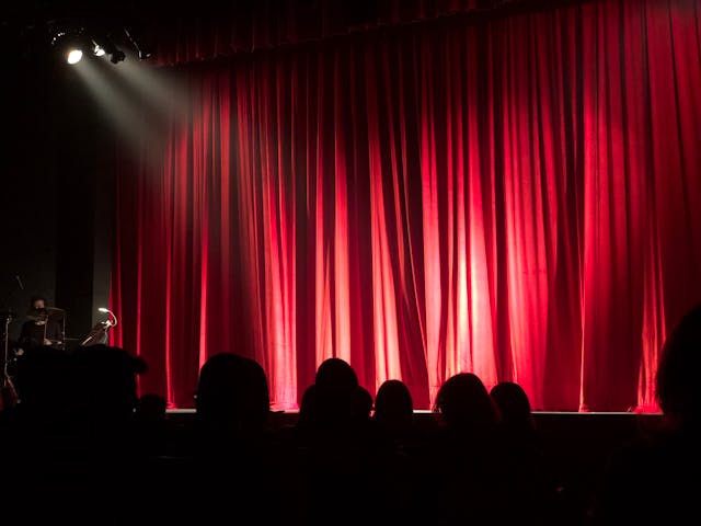 An audience sits and waits for the red curtains on the stage to open.