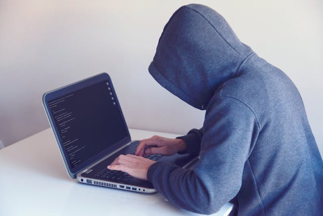 匿名の人物がフード付きのセーターを着て、ノートパソコンで文字を打っている。