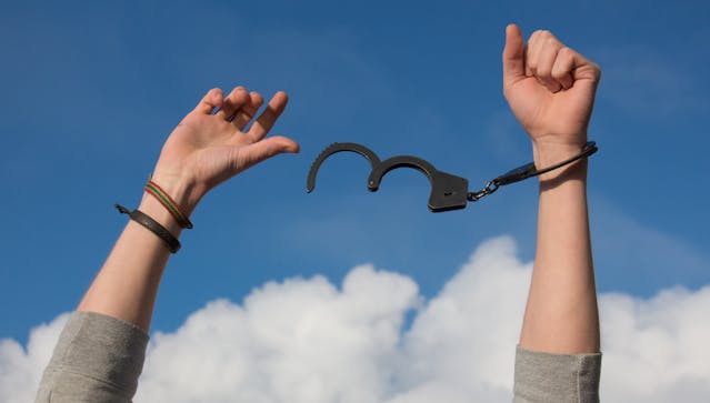 A pair of handcuffs dangles from one arm as the person holds both arms up to the sky.