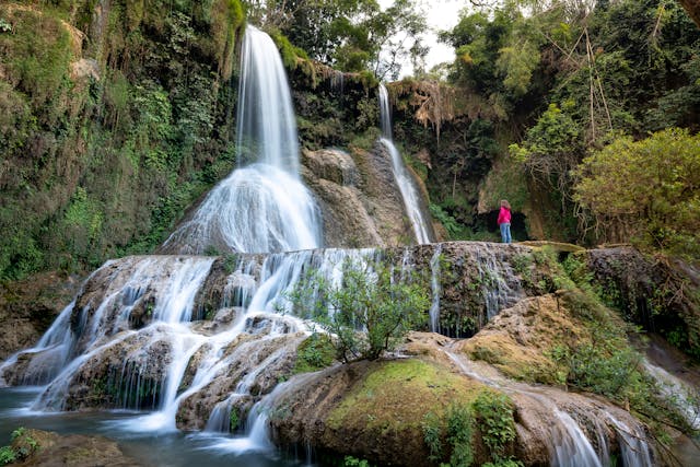 A woman stands in front of towering waterfalls.