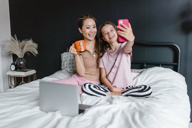 Uma mãe e sua filha sorrindo e sentadas em uma cama enquanto tiram uma selfie com um smartphone rosa.