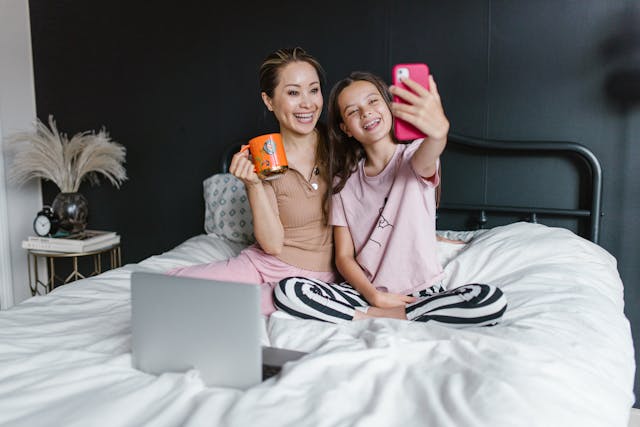 Een moeder en haar dochter glimlachen en zitten in een bed terwijl ze een selfie nemen met een roze smartphone.