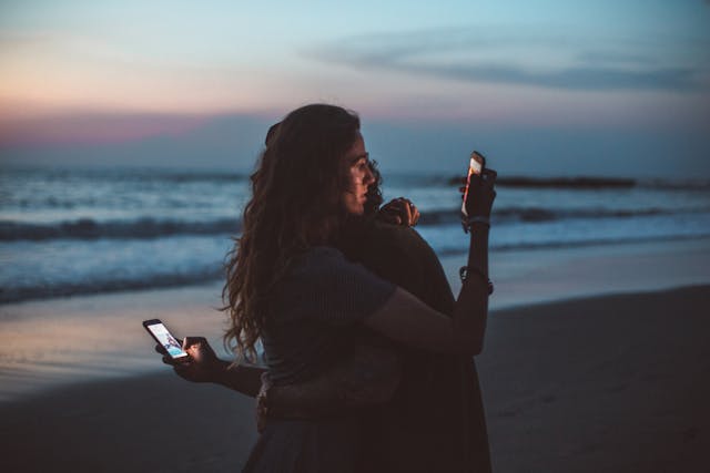 A couple at the beach hugs each other while they also look at their phones. 
