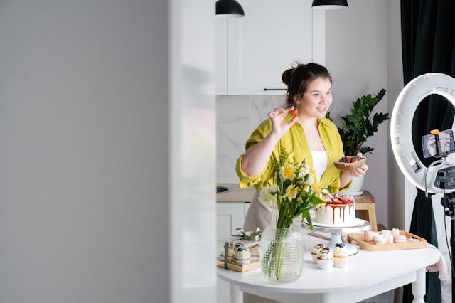 Uma mulher está em frente a uma mesa cheia de bolos e doces enquanto realiza uma transmissão AO VIVO no TikTok. 