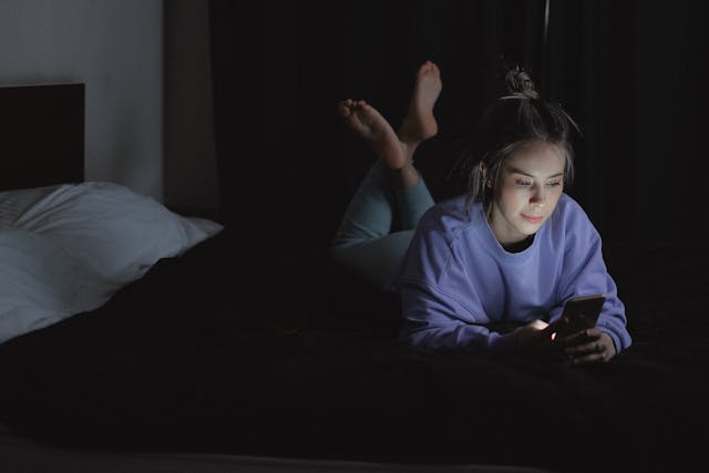 暗い寝室でうつぶせになり、携帯電話でビデオを見ている女性。 