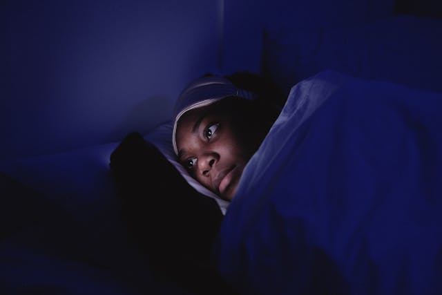 Une femme avec un masque de sommeil sur la tête est allongée dans son lit et navigue sur son téléphone dans l'obscurité.