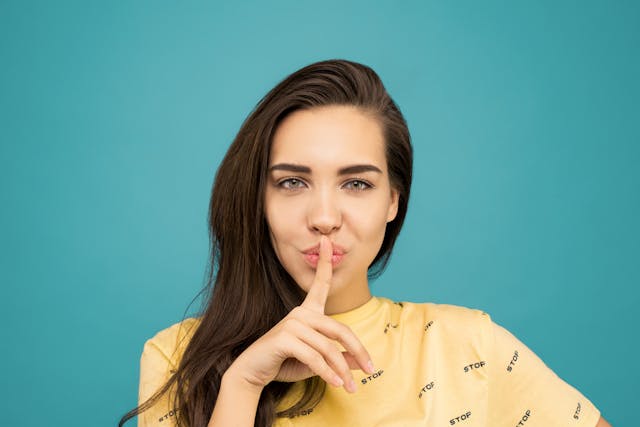 A woman stands in front of a blue wall and makes the shush sign with her index finger on her lips. 
