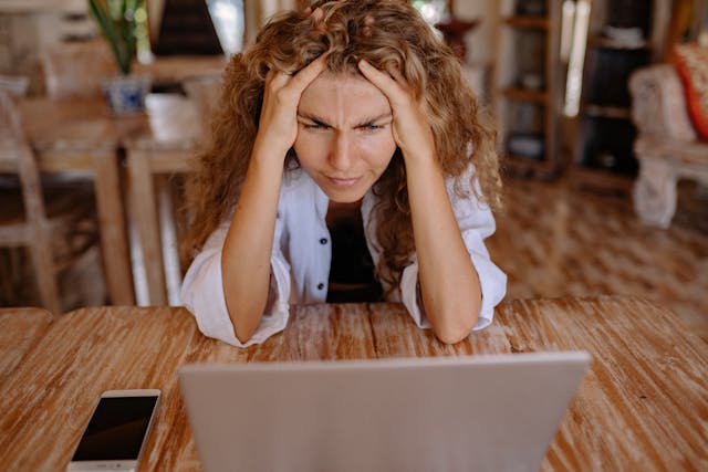 A woman has both hands in her hair as she stares confusedly at her laptop.