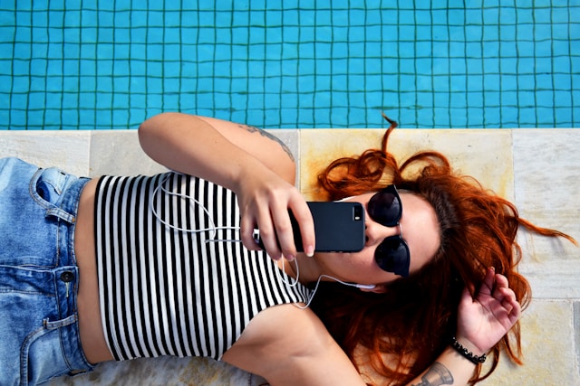 A woman with earphones lies down beside a pool and watches TikToks on her mobile device.