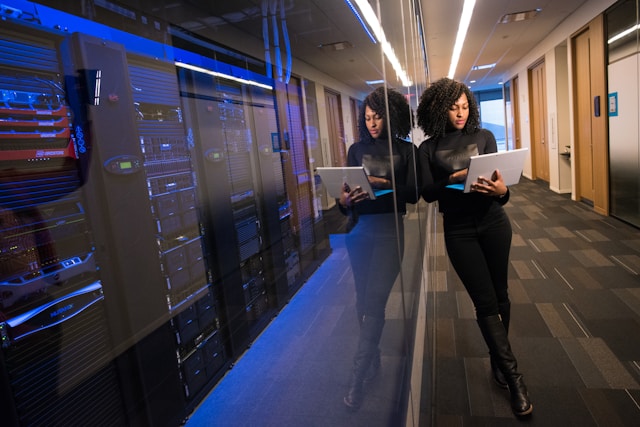 A person uses their laptop while standing beside a server room.
