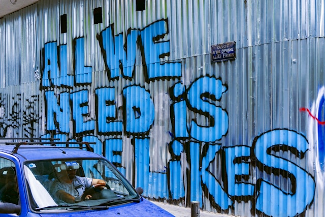 Blue graffiti on a wall reads, “All we need is more likes.”

