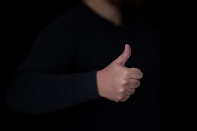 Una persona con una camicia nera a maniche lunghe fa il segno di ok con il pollice alzato. 