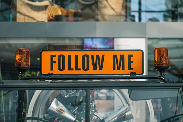 Uma placa preta e laranja diz: "Follow me" (Siga-me).